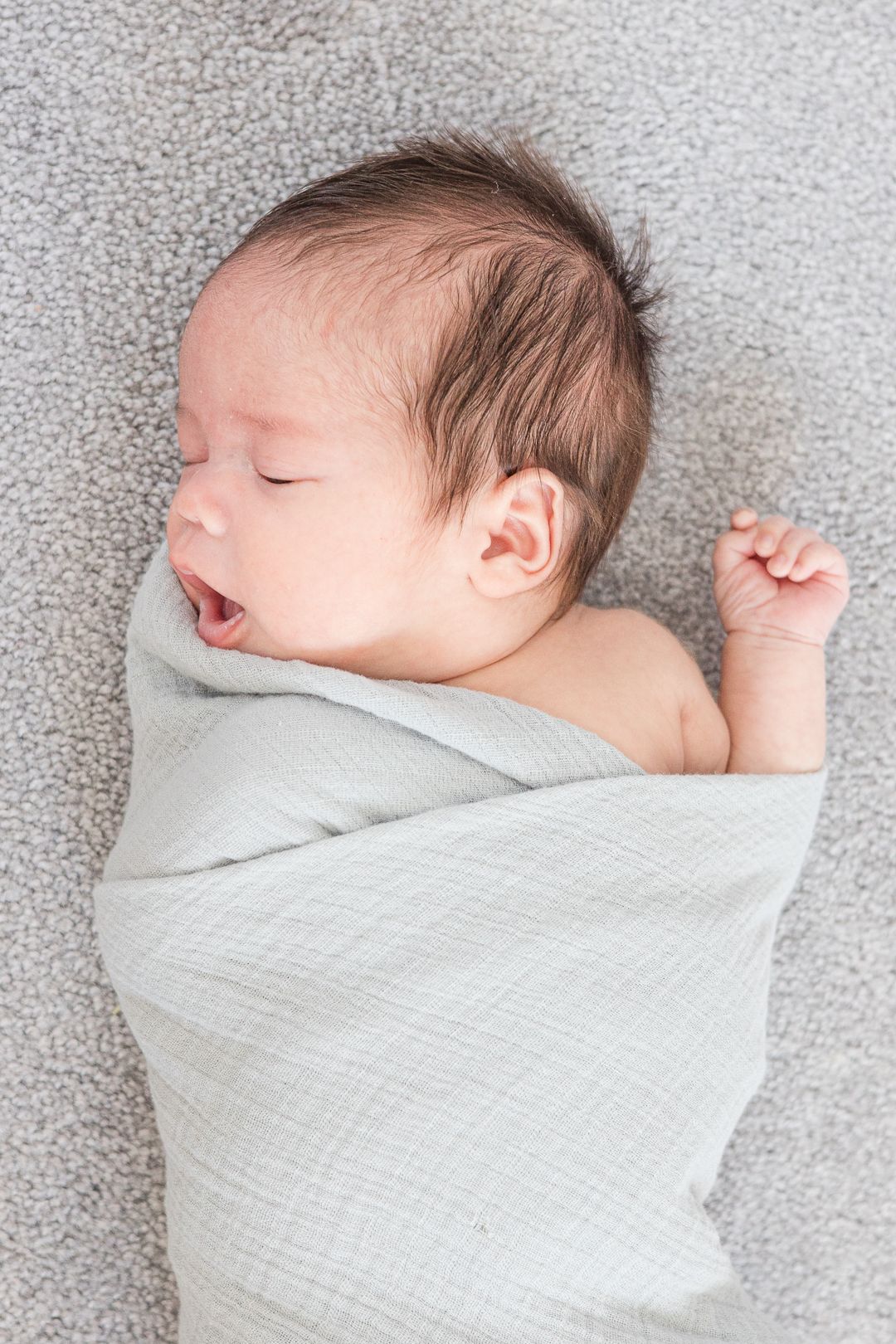 A sleeping newborn baby swaddled in light gray fabric lies on a gray textured surface.