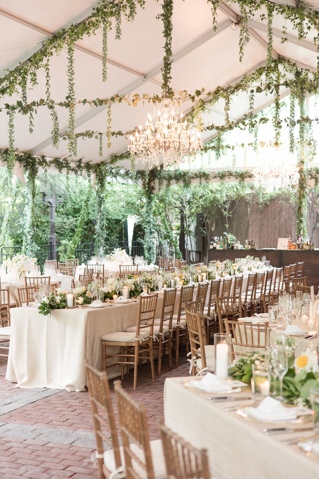 Romantic wedding reception setup with crystal chandelier and cascading greenery in an outdoor tent.