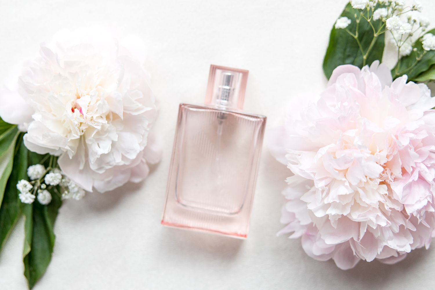 Pink perfume bottle styled with white peonies and green leaves on white surface.