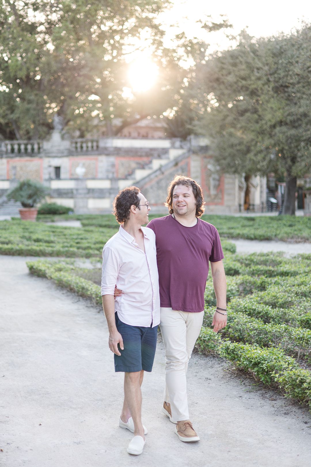 A couple walks along a garden path at sunset near a historic building.
