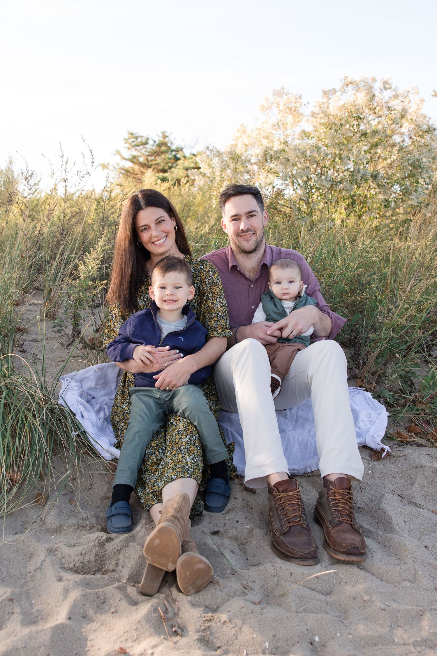 Family sitting together on a sandy beach blanket surrounded by tall beach grass.