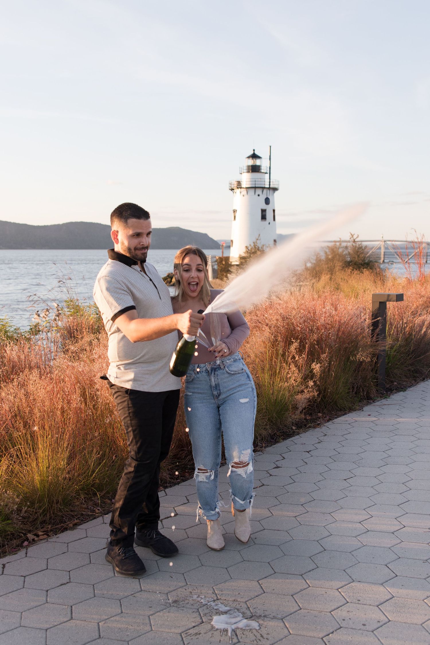 A couple celebrates with champagne near a lighthouse at sunset on a stone pathway.