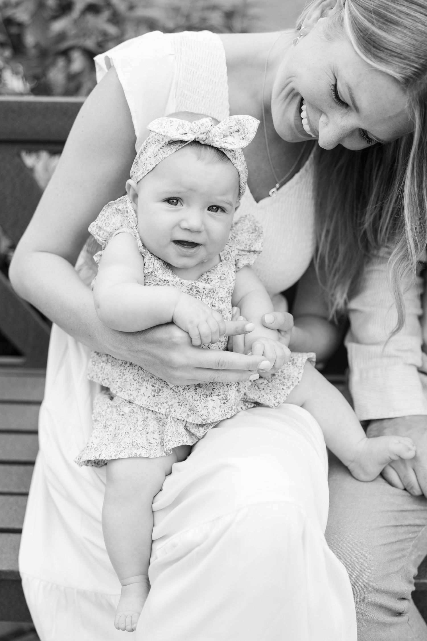 A sweet black and white portrait of a parent holding a baby in a lace dress and headband sitting outdoors.