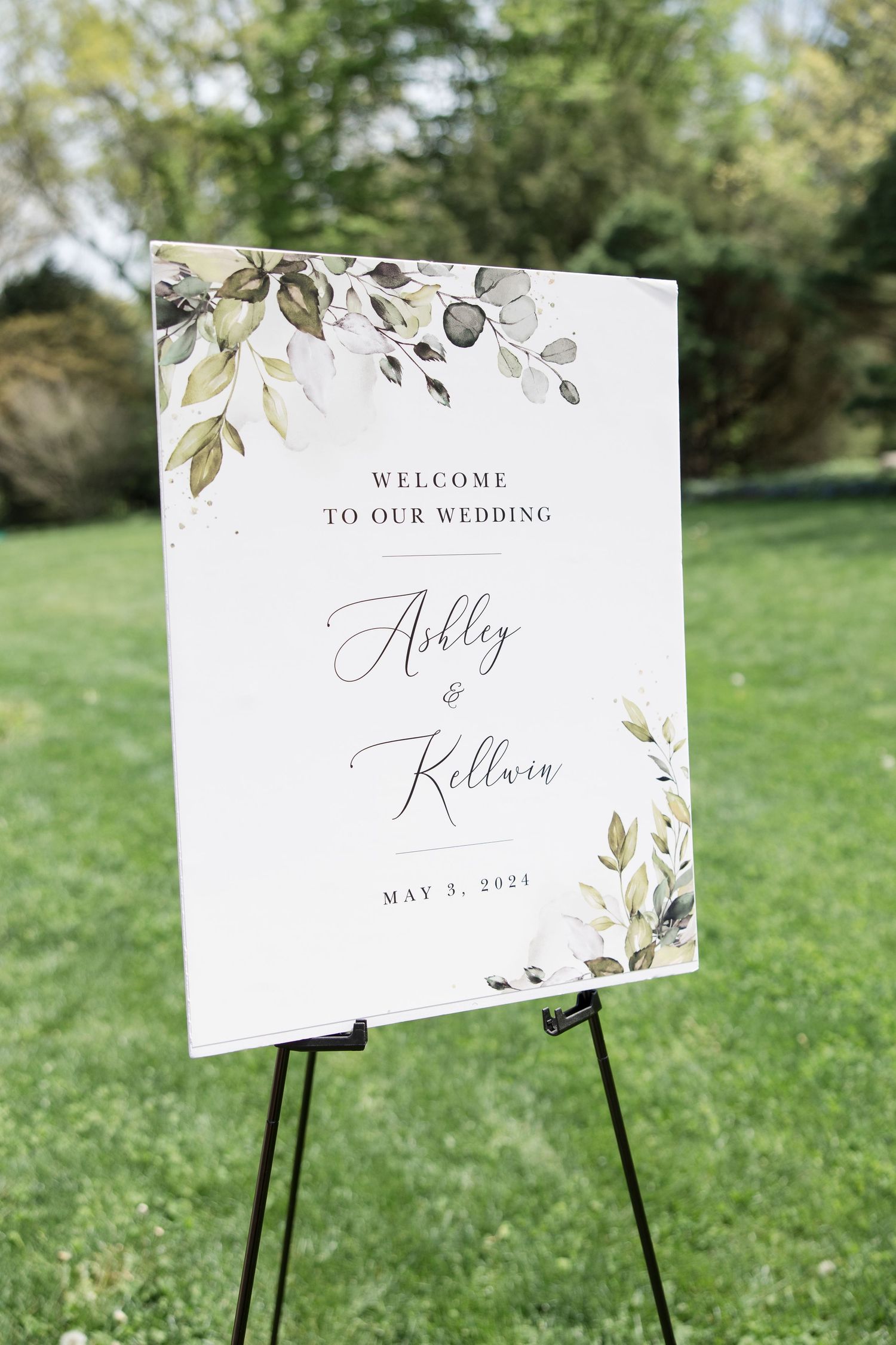 Elegant wedding welcome sign with watercolor floral design displayed on black easel outdoors.