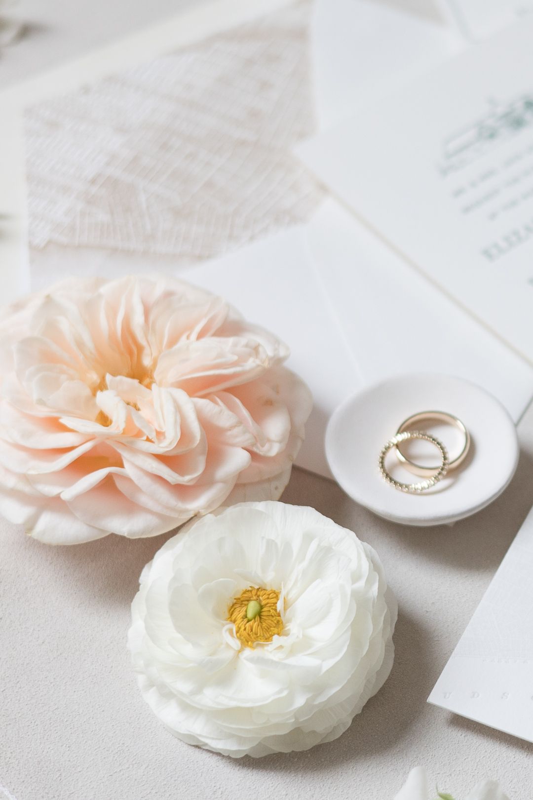 Wedding rings nestled in a white ceramic dish with pink and white flowers on a light table.