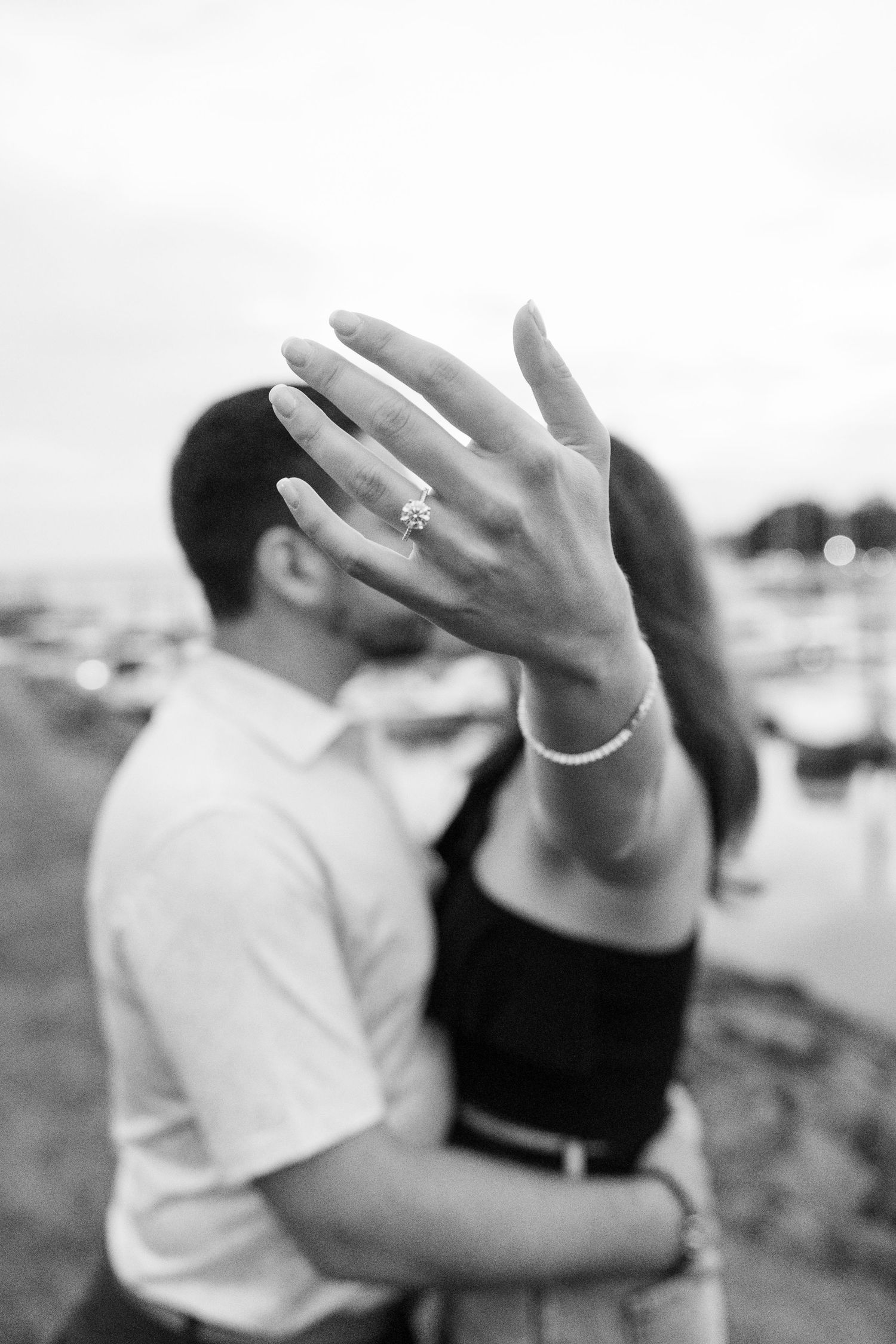 A couple sharing an intimate moment as a hand with an engagement ring is held up in black and white.