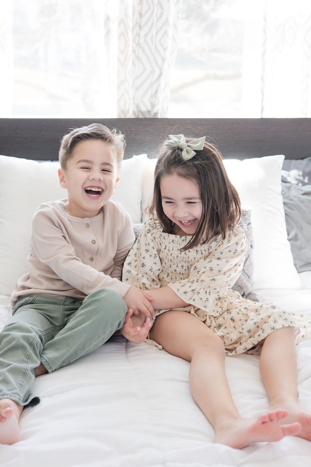 Two siblings share a joyful laugh while sitting on white bedding.