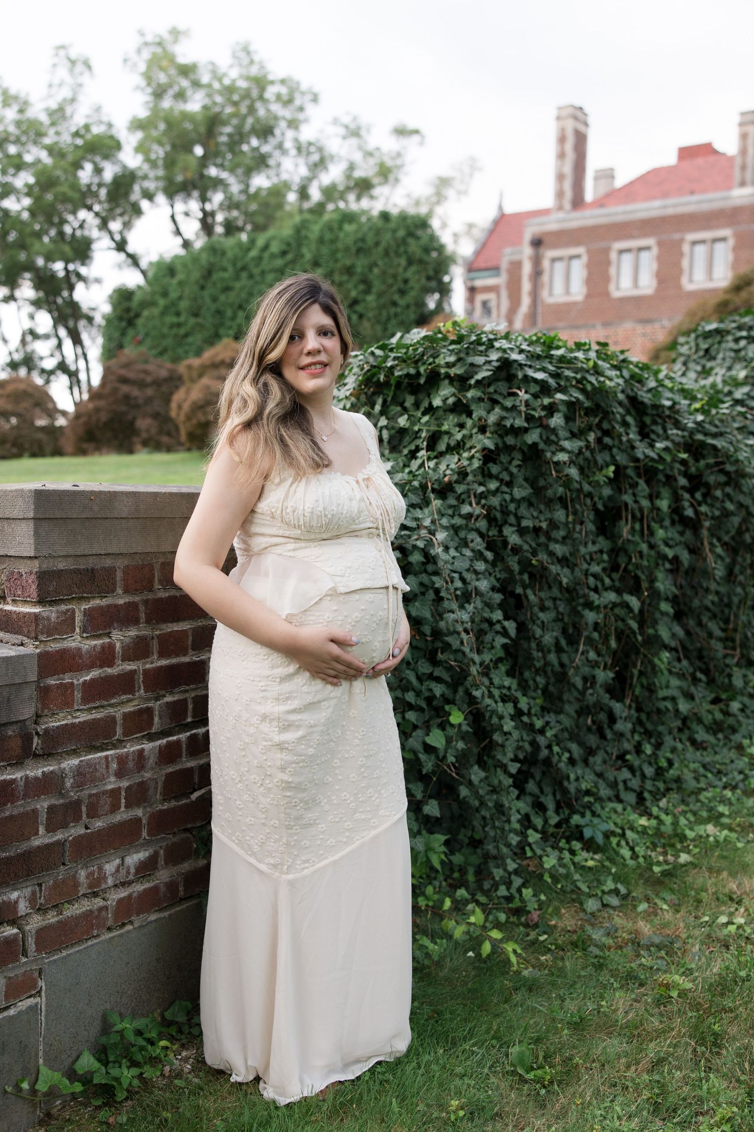 Pregnant woman in flowing white dress stands beside brick wall and greenery.