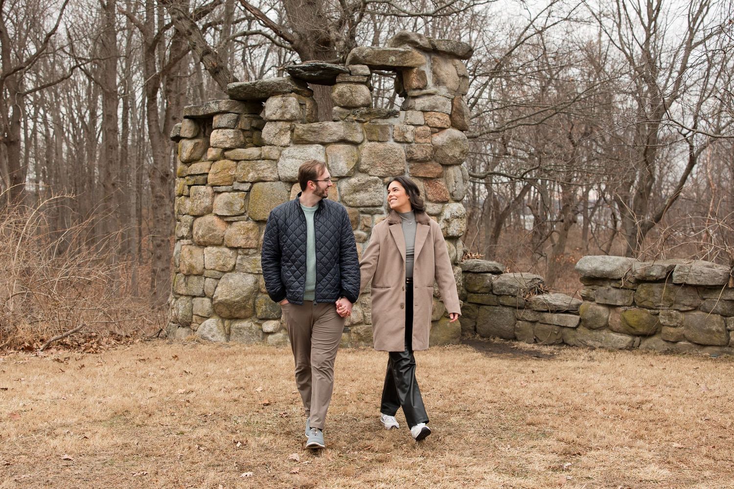 A couple walking hand in hand near a rustic stone wall structure in a park.