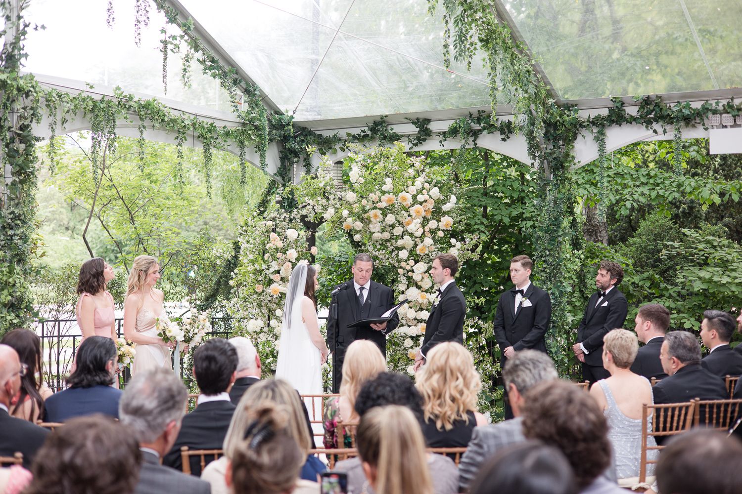 A sunlit outdoor wedding ceremony with guests seated in front of a floral-adorned arch in a lush garden setting.