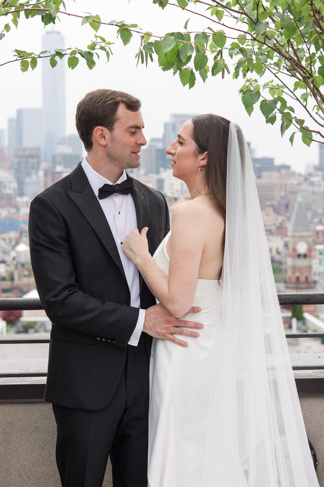 Romantic embrace on a Manhattan rooftop terrace with city views and leafy branches overhead.