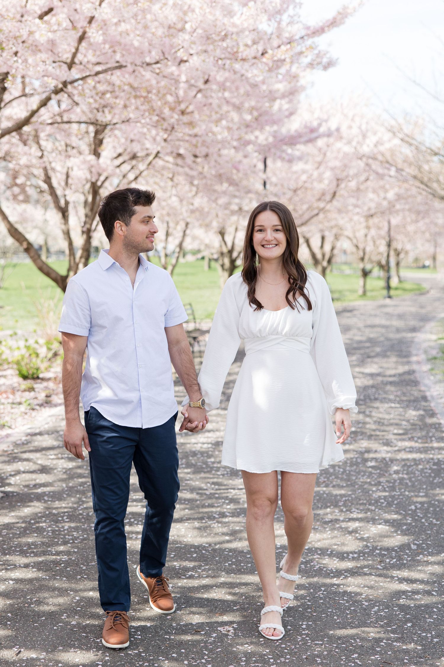 Couple walking hand in hand down path lined with blooming cherry blossom trees.