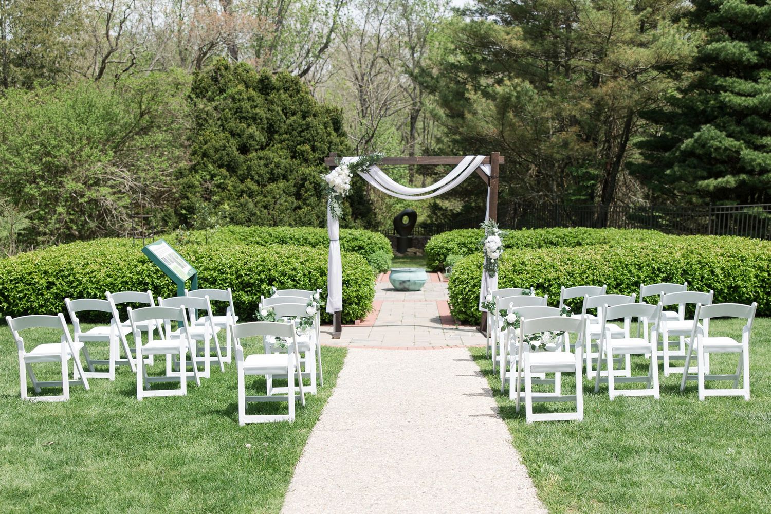 Intimate outdoor wedding venue with white chairs lined along stone path leading to simple ceremony arch.