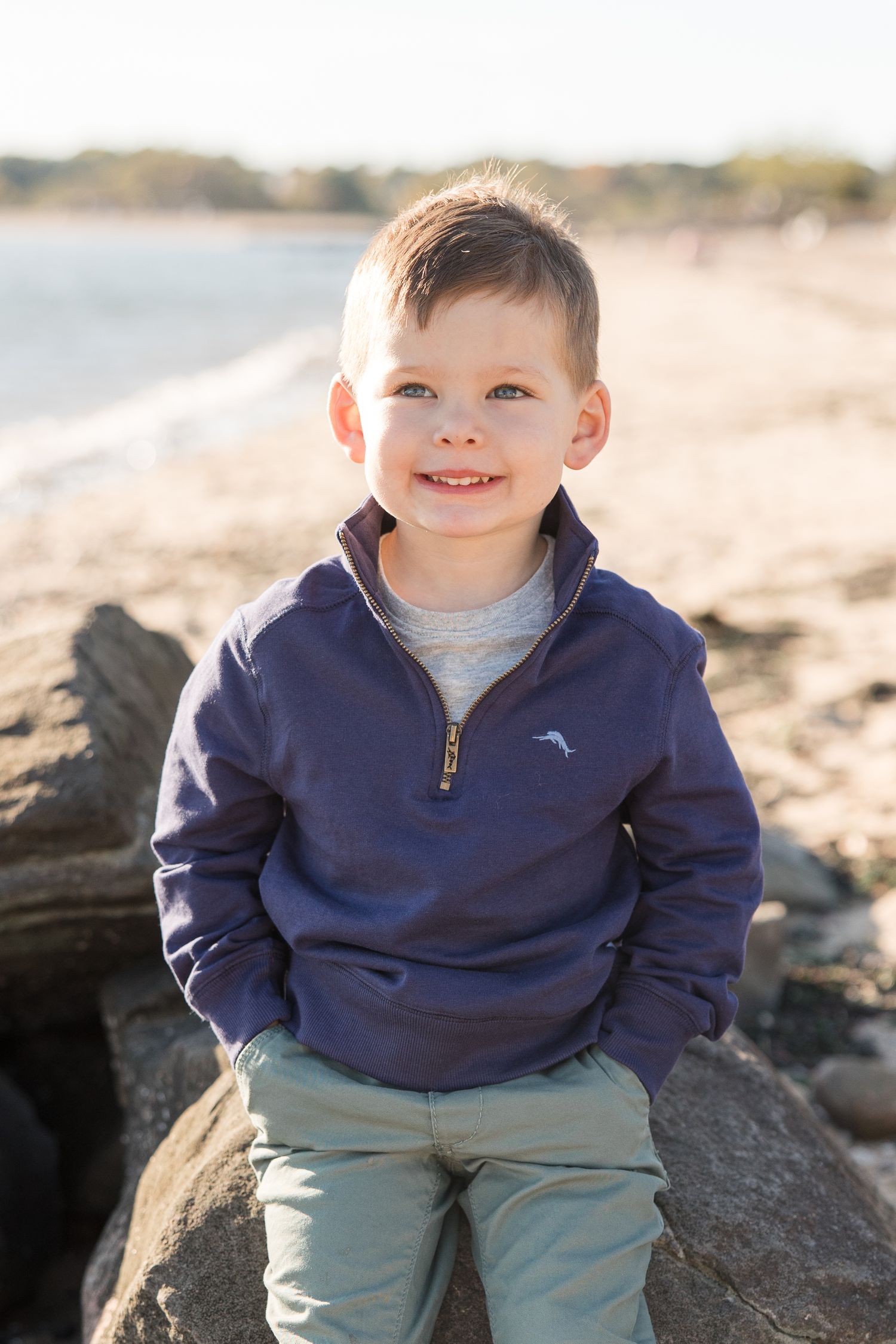 Young child in navy quarter-zip sweater sitting on beach rocks.