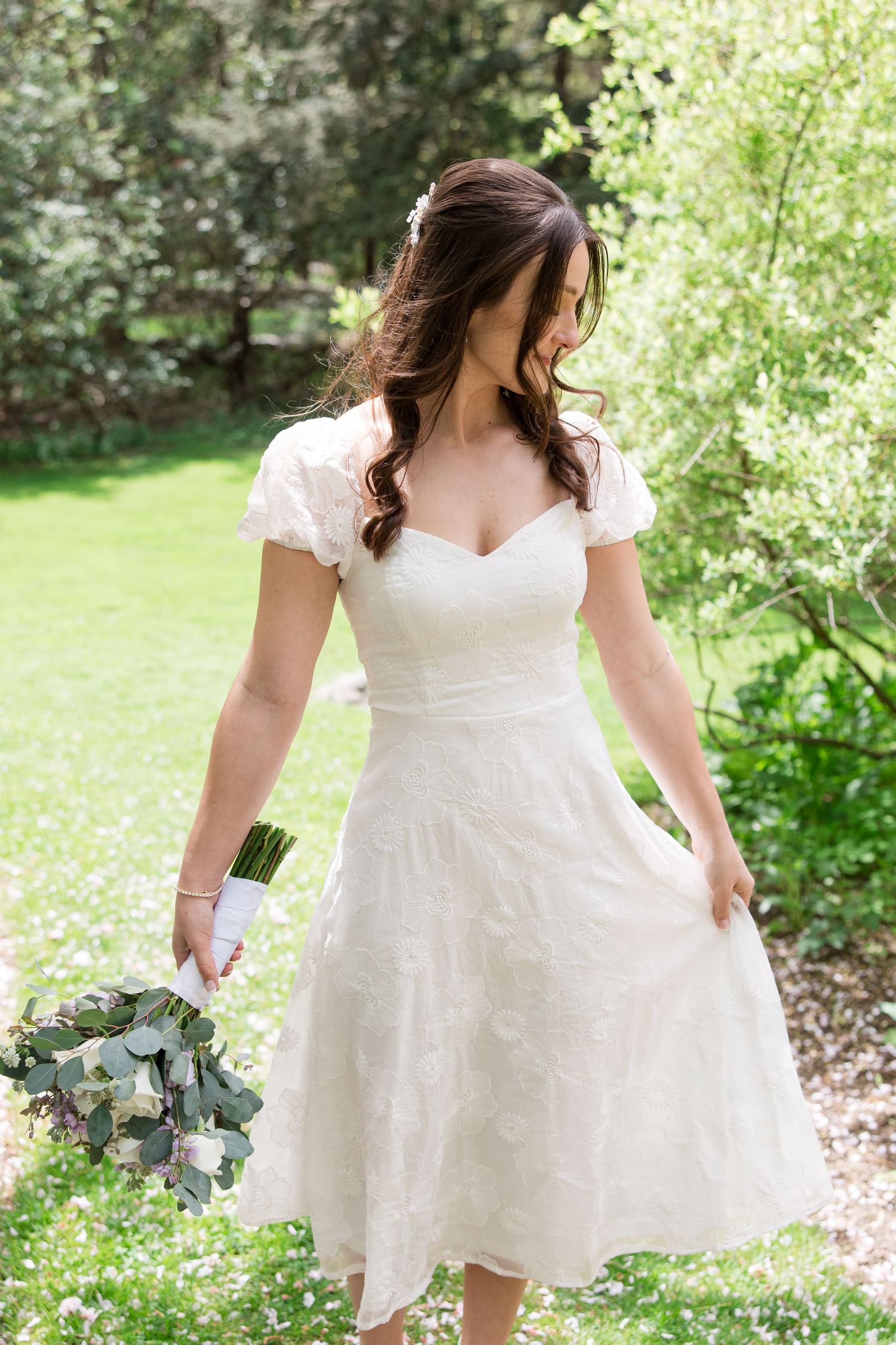 A bride in a tea-length white lace dress with cap sleeves stands in a garden setting holding a eucalyptus bouquet.