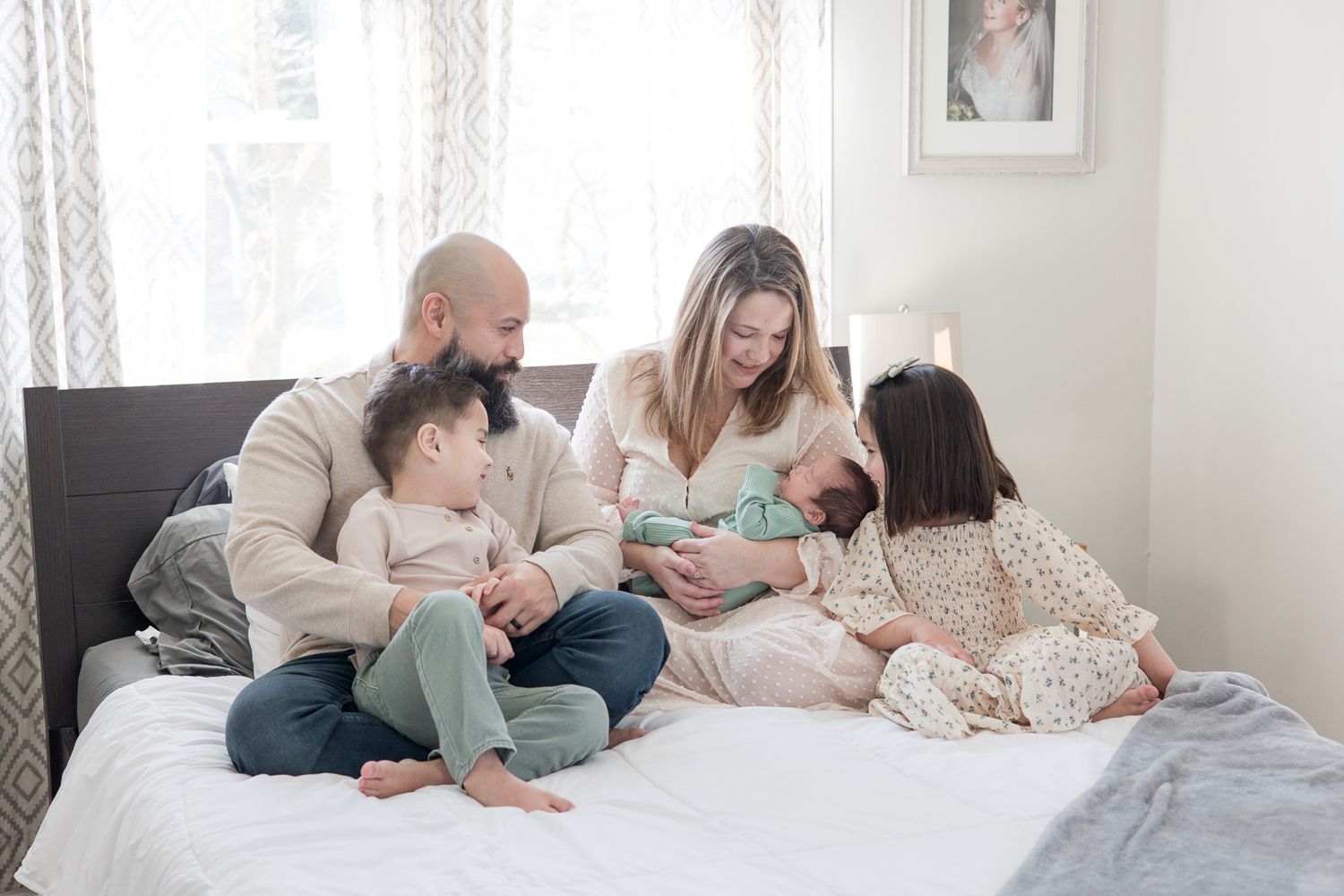 Family lounges together on white bedding wearing casual clothing in soft neutral tones.