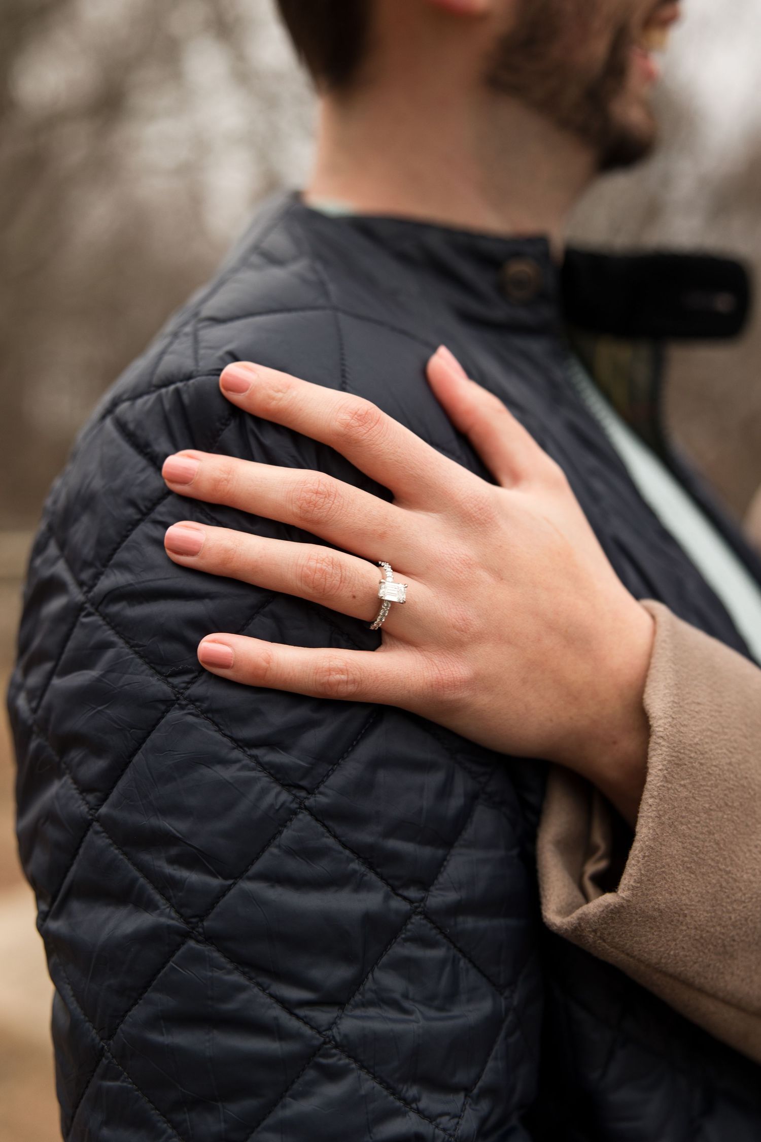 A close-up shot of a hand wearing an engagement ring against a quilted jacket.