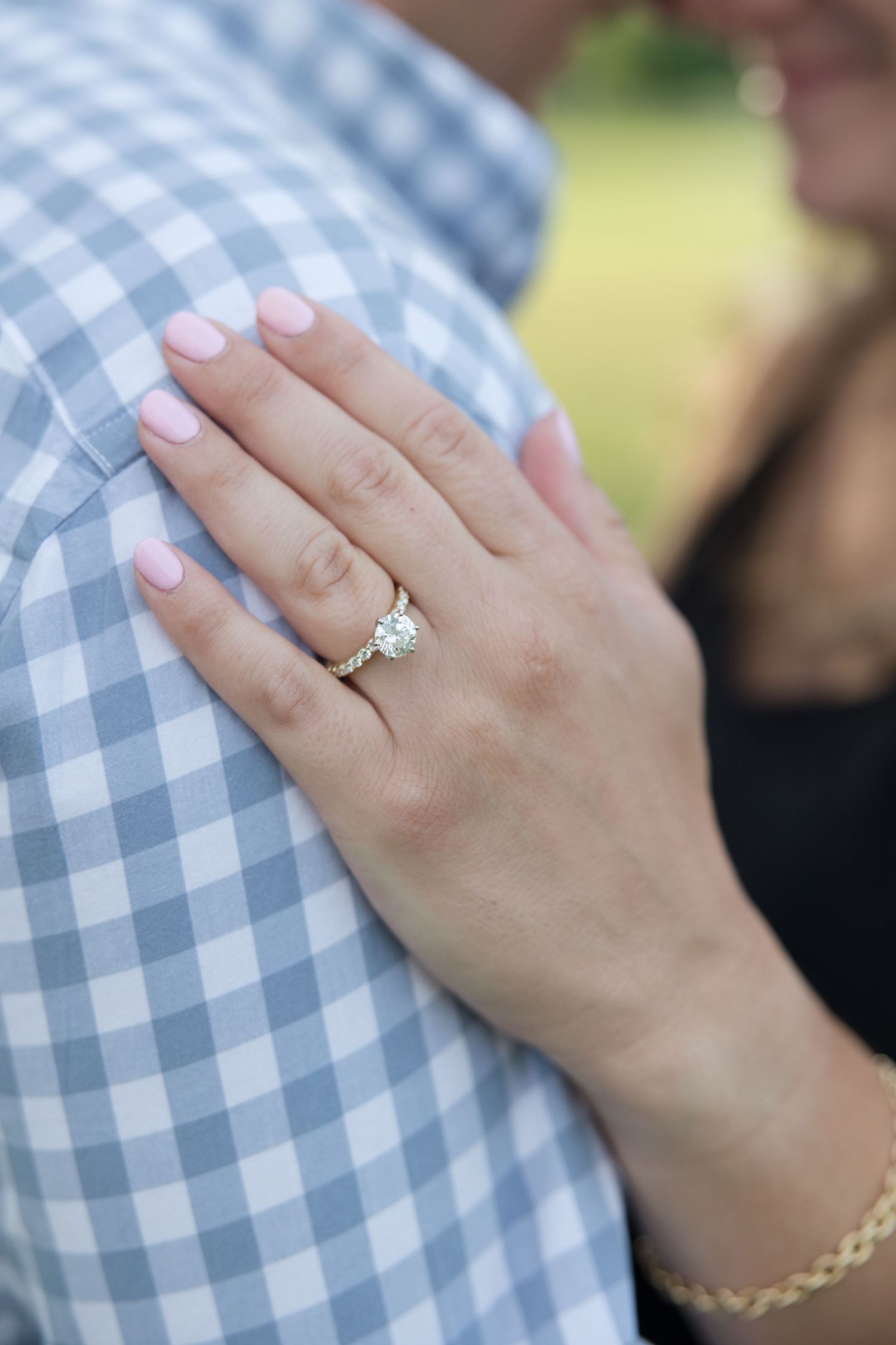 Close-up of a diamond engagement ring on a hand resting against a blue gingham shirt.
