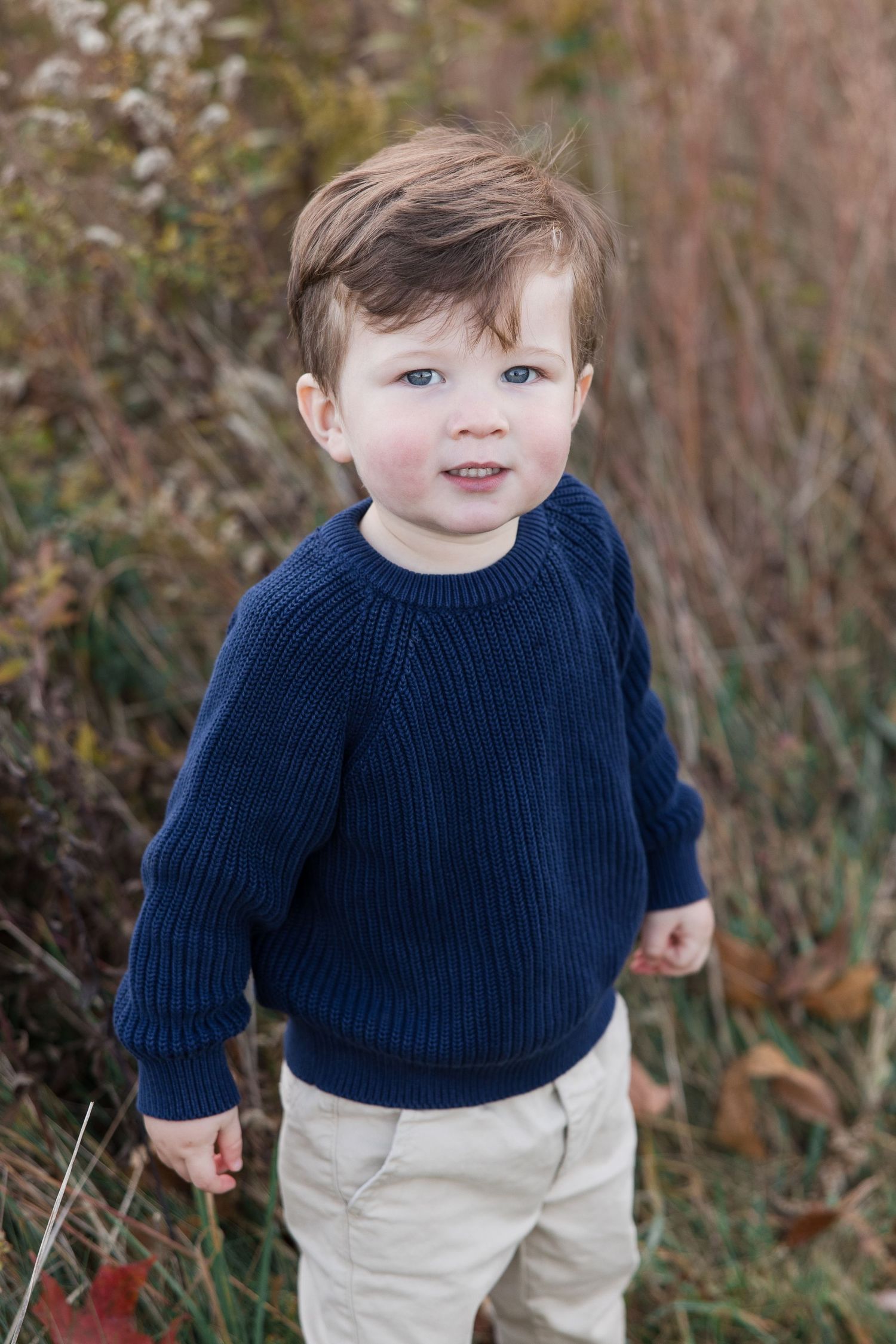 A toddler in a navy sweater and khaki pants smiles while standing outdoors.