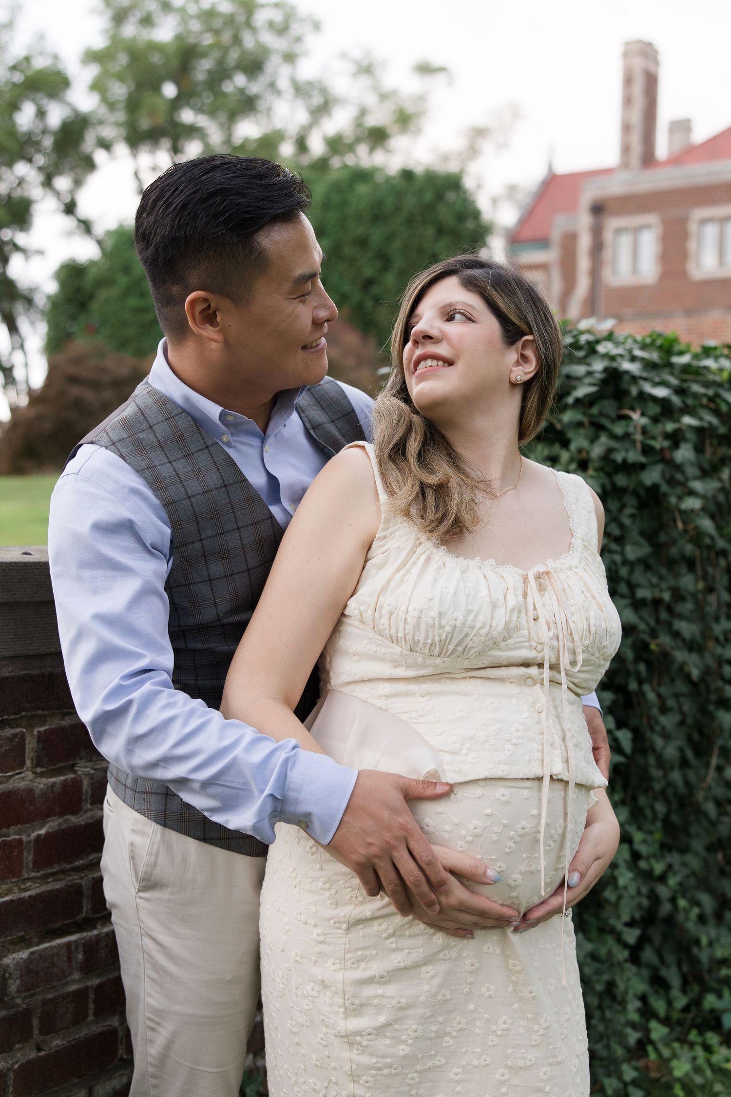 Expecting couple embraces outdoors near a brick wall and ivy-covered building.