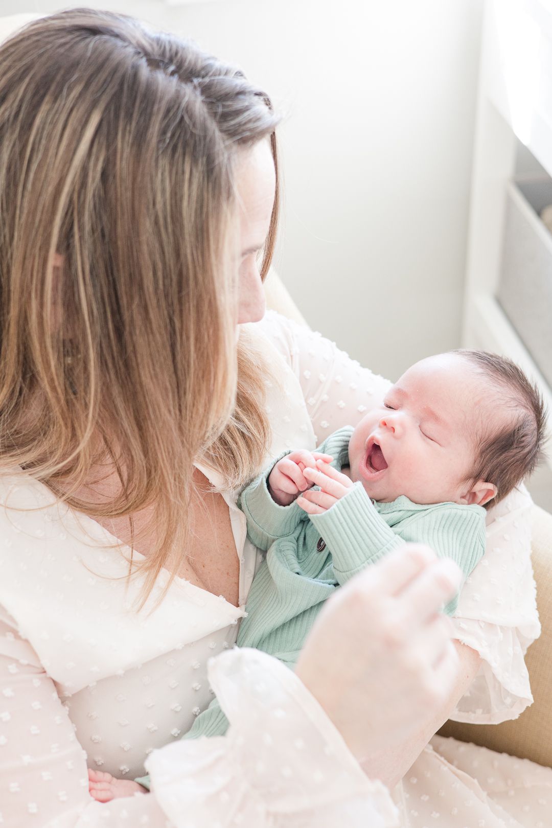 A woman in white holds a yawning baby wearing mint green while sitting in a cream armchair.