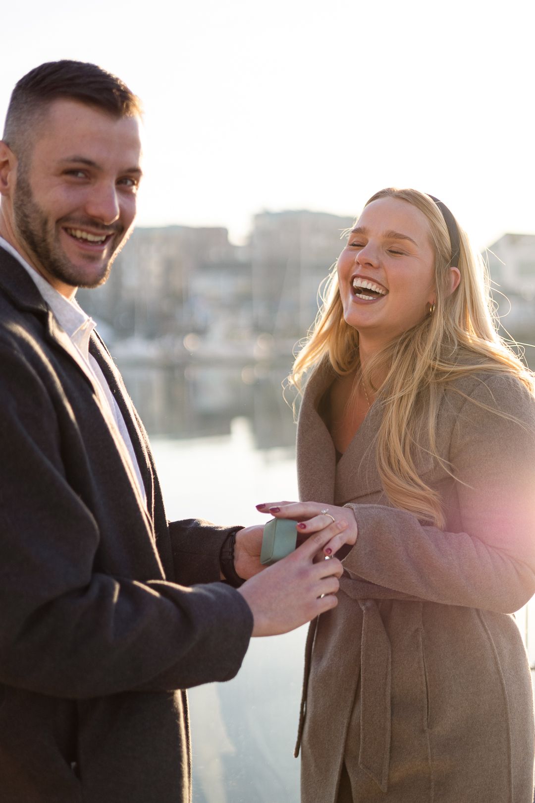 Joyful reaction during waterfront proposal with marina and boats in background.