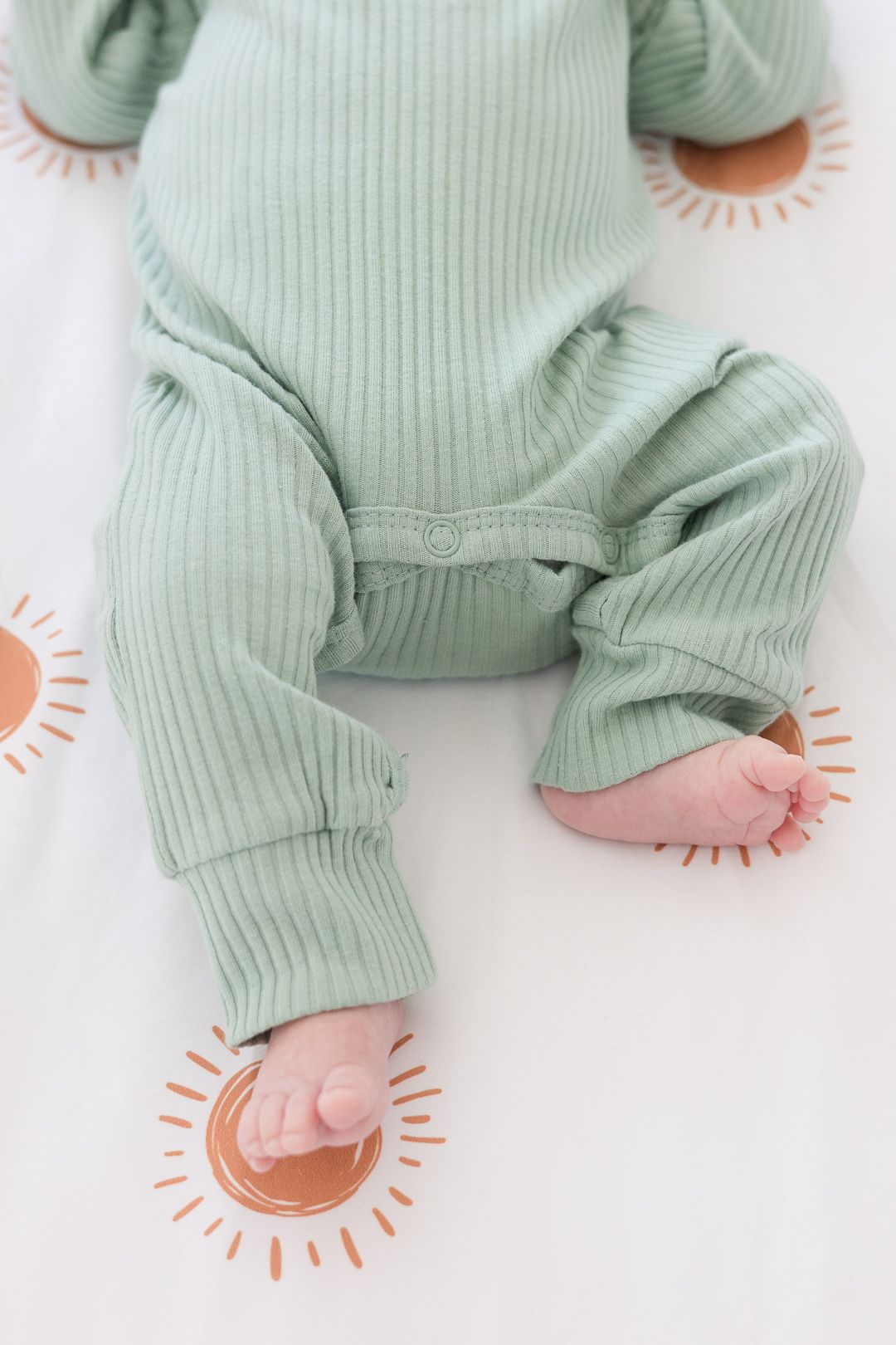 Baby wearing mint green ribbed onesie lying on white fabric with peach sun pattern.
