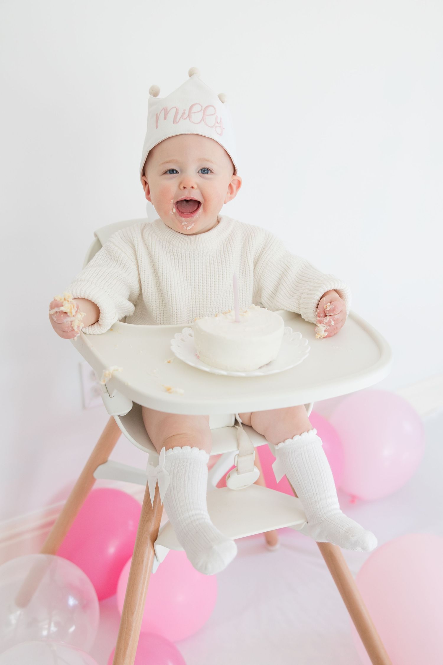 A smiling baby wearing a white sweater and crown sits in a high chair with pink balloons in the background.