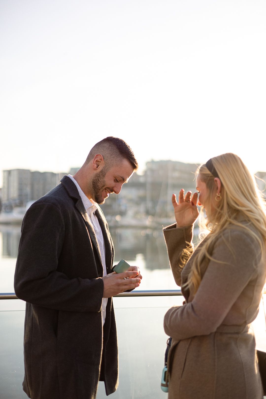 Candid engagement moment captured on marina dock with city buildings in background.