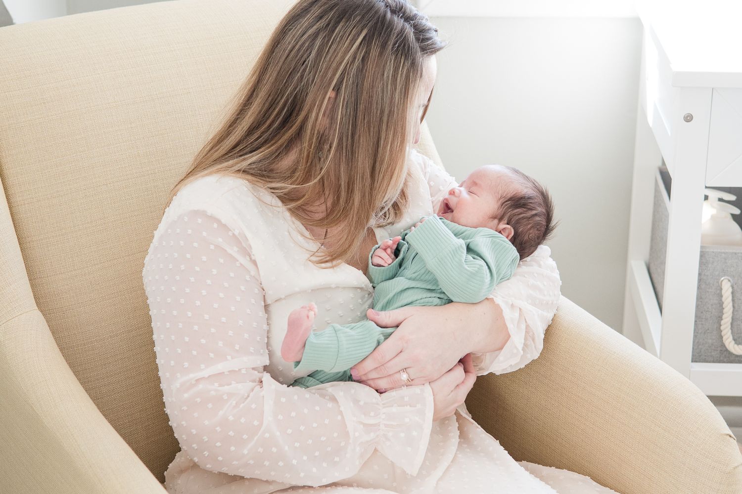 A woman in a dotted white dress gazes lovingly at a newborn baby dressed in mint green.