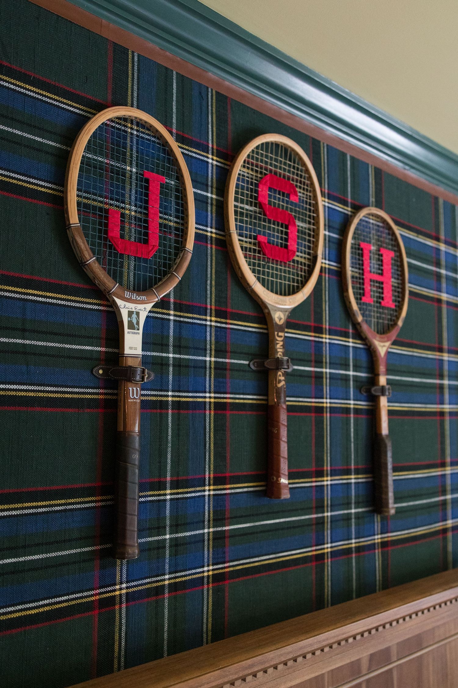Three vintage tennis rackets with red letters JSH mounted on plaid wallpaper.