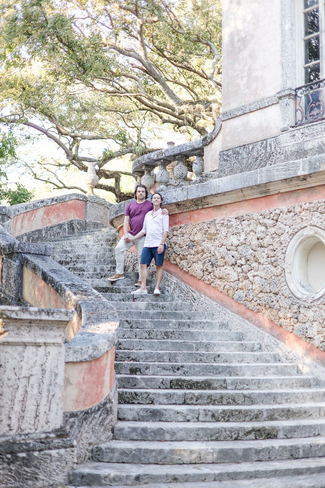 Two people pose on grand stone steps of a historic building with overhanging trees.
