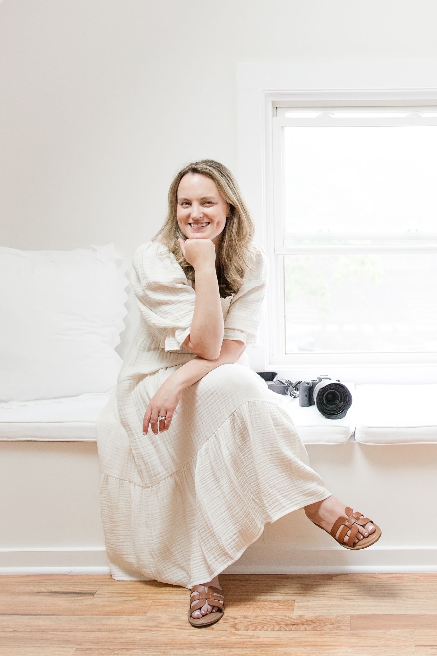 Person in white summer dress sits on windowsill with natural light and camera nearby in minimal white room.