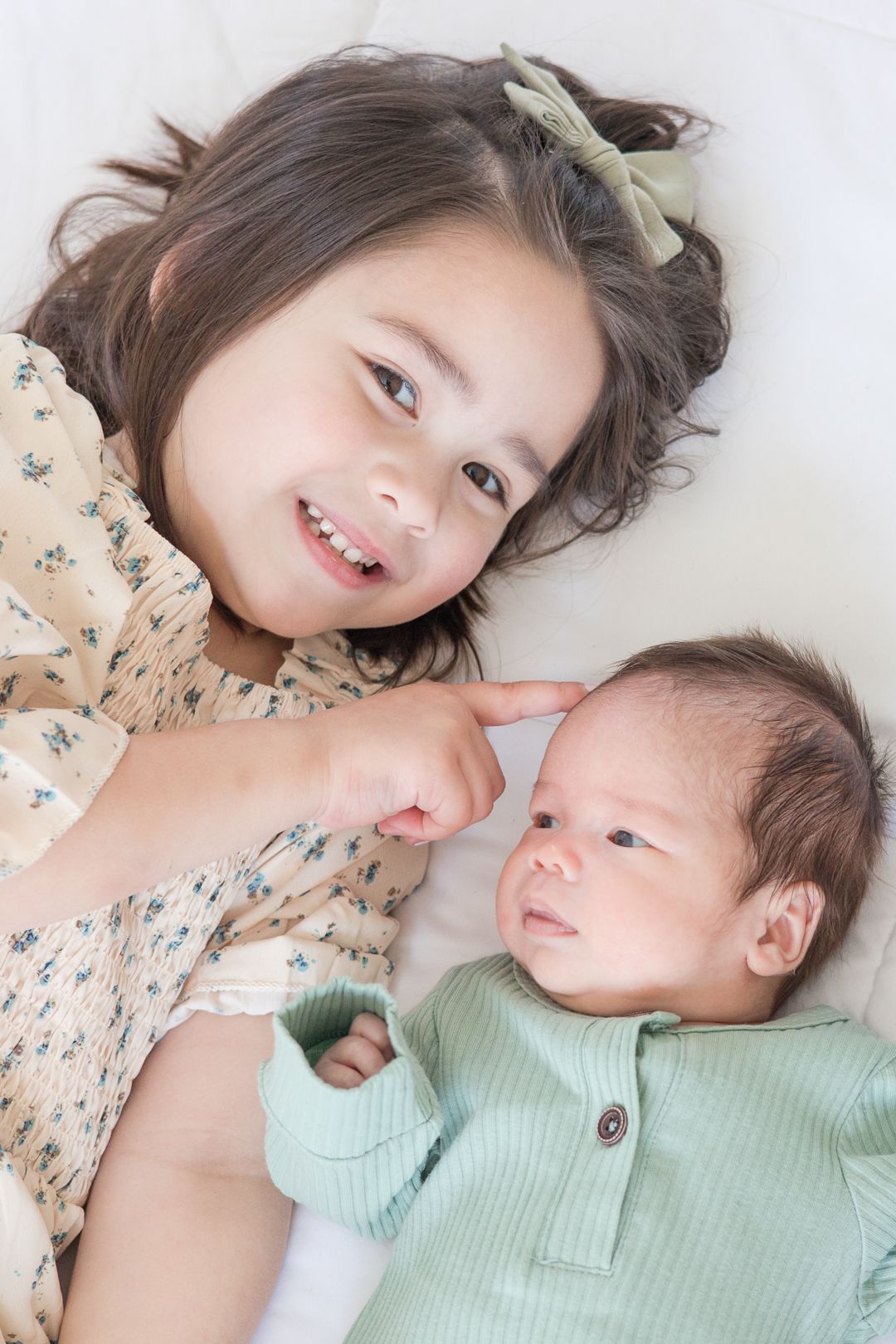 Two siblings share a sweet moment lying together on white bedding with the baby in mint green.