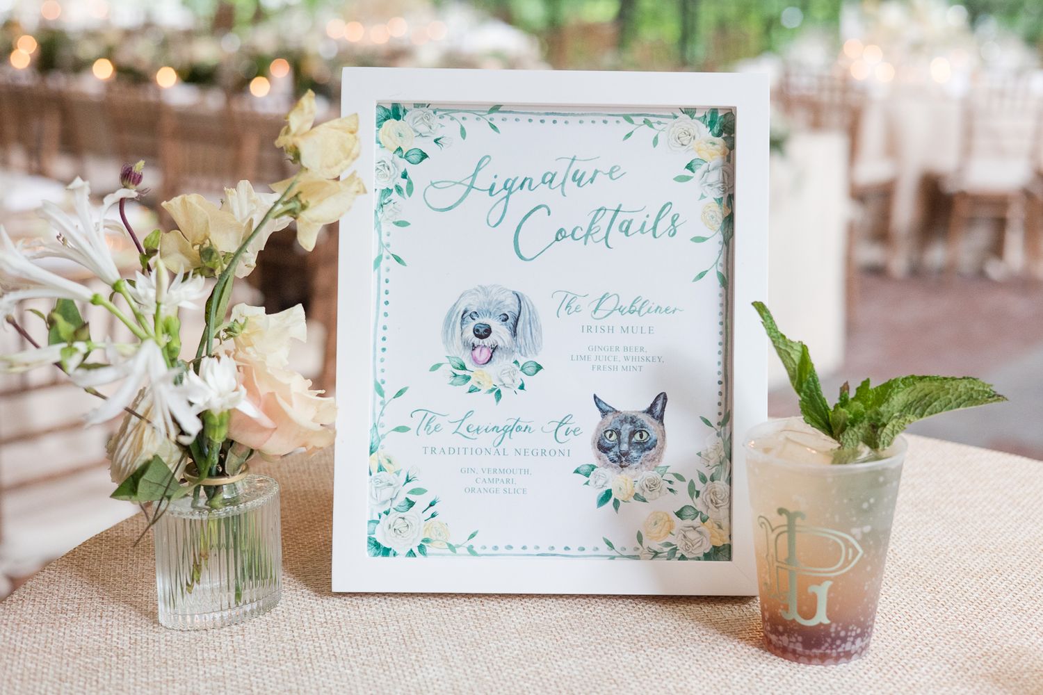 Elegant wedding signage with illustrated pets and floral design in a white frame next to delicate flower arrangements.