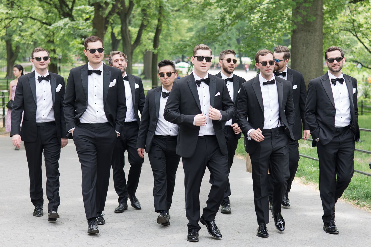 Groomsmen in black tuxedos and sunglasses walking together on a tree-lined path.