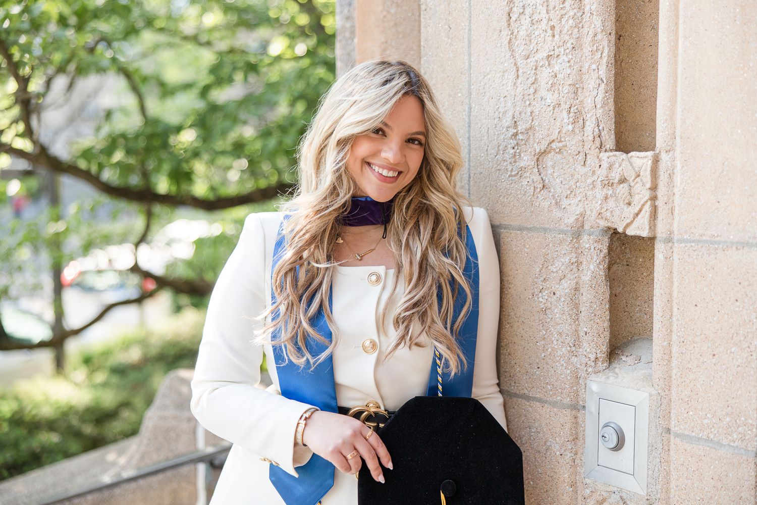 Professional portrait against stone building wearing white blazer and blue vest.