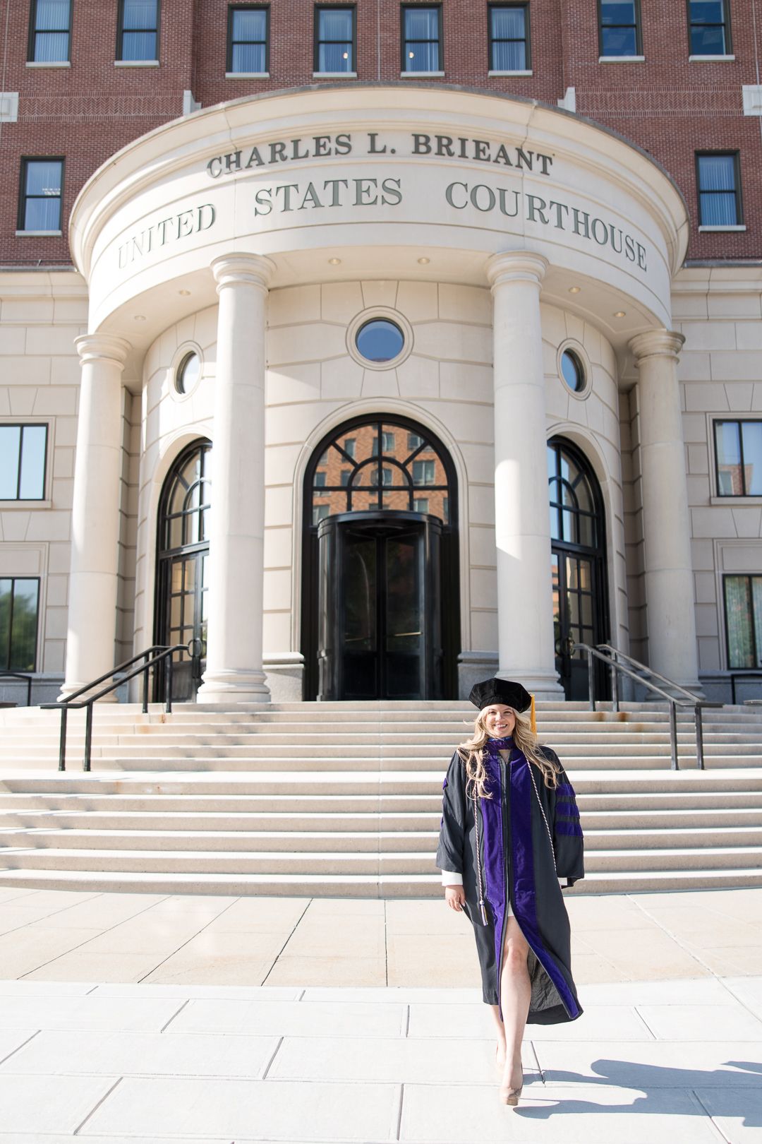 Graduate in purple regalia standing on courthouse steps in front of classical architecture.