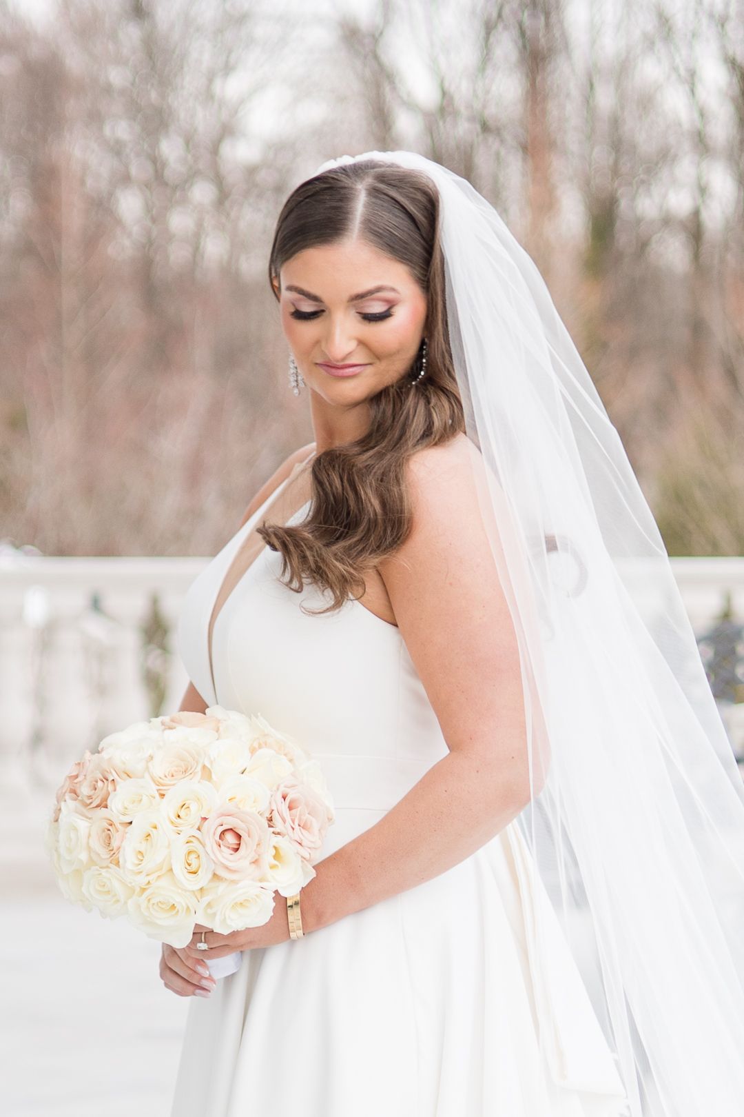 A bride in a white wedding dress holding a pink and cream rose bouquet with flowing veil.