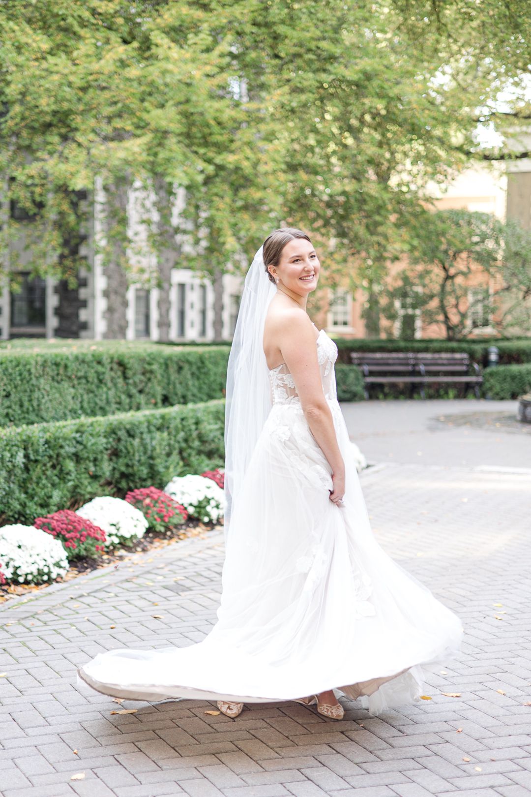 A bride in a flowing white dress twirls on a brick courtyard surrounded by trees.