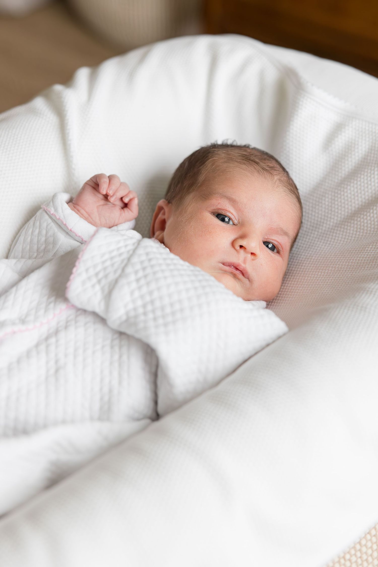 Baby in white outfit lying on white cushioned surface looking at camera.