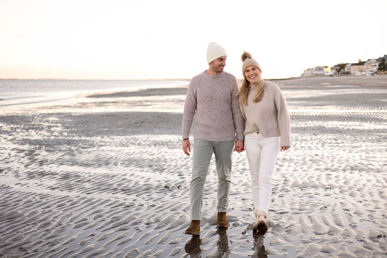 Couple in matching neutral sweaters walking along beach at sunset.