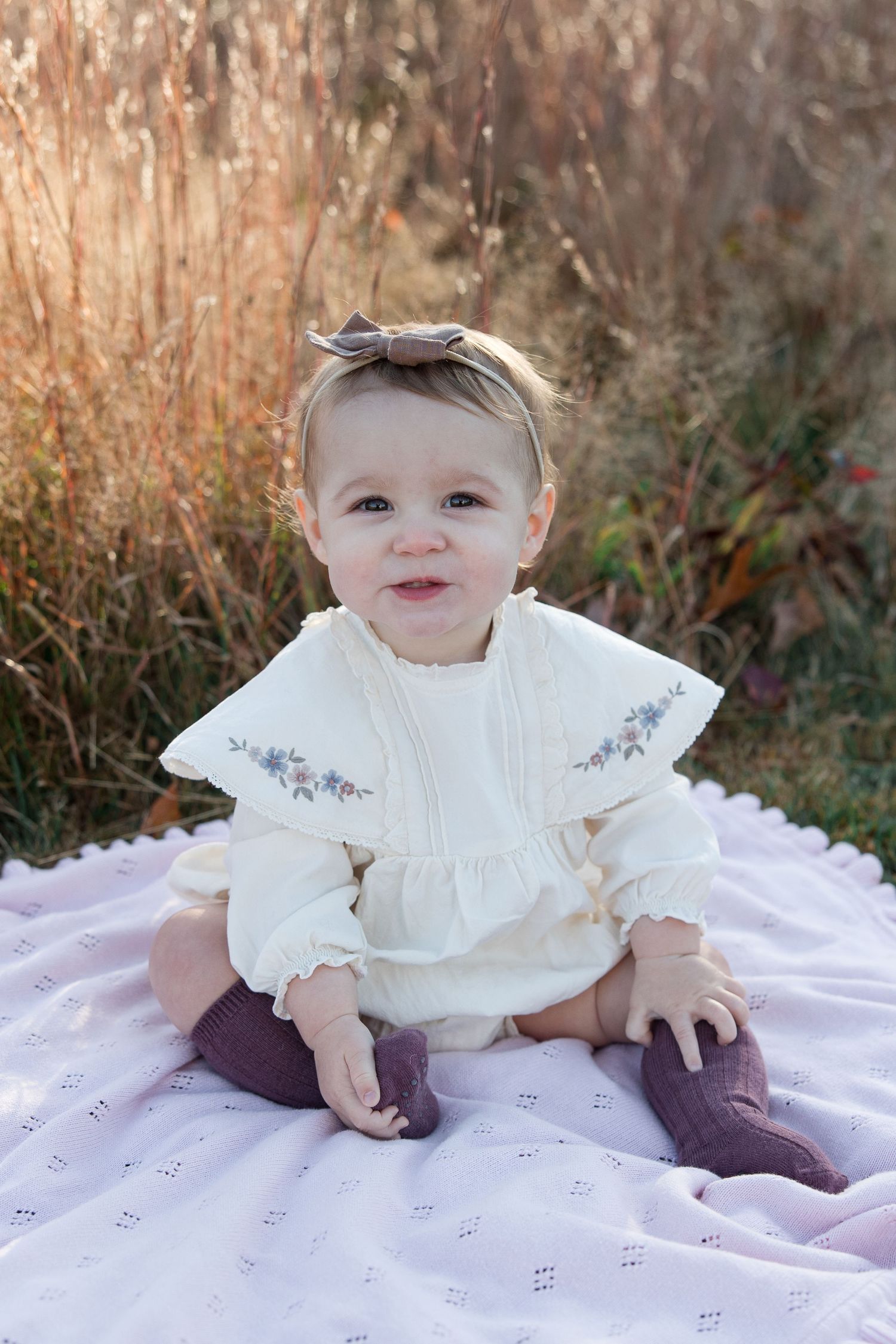 Smiling baby in white dress with bow headband sitting on purple blanket outdoors.