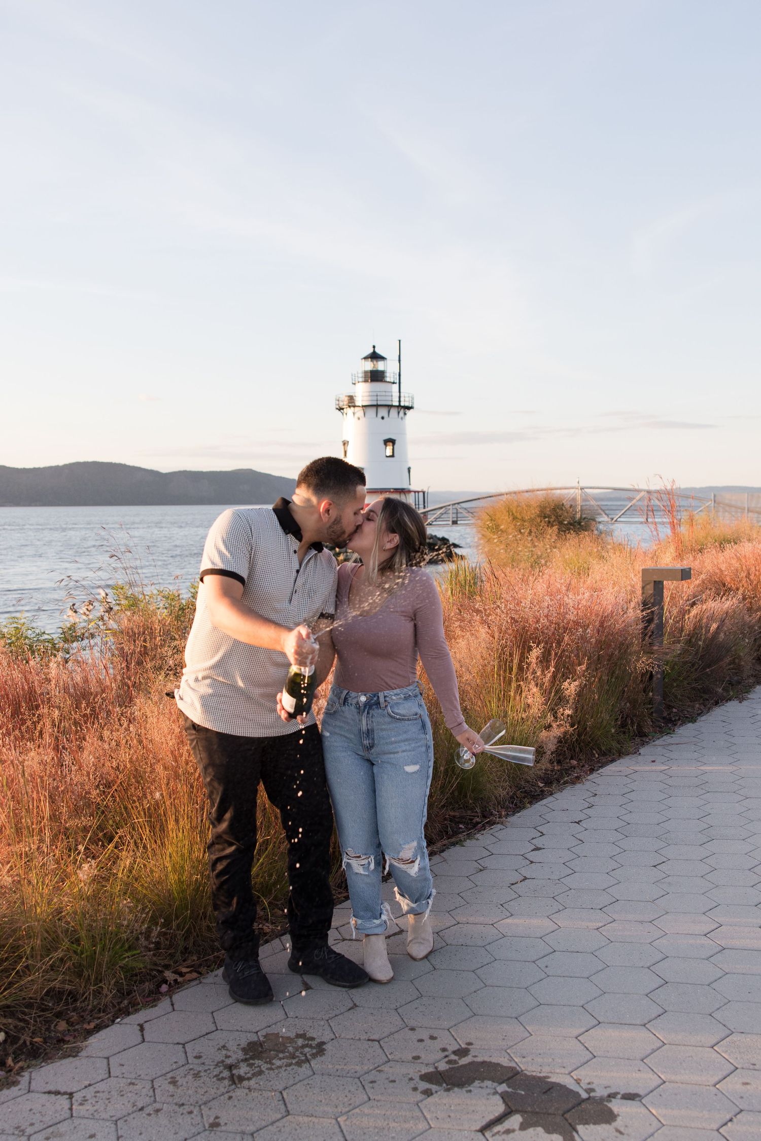 A couple sharing a romantic moment on a coastal path with a lighthouse in the background.