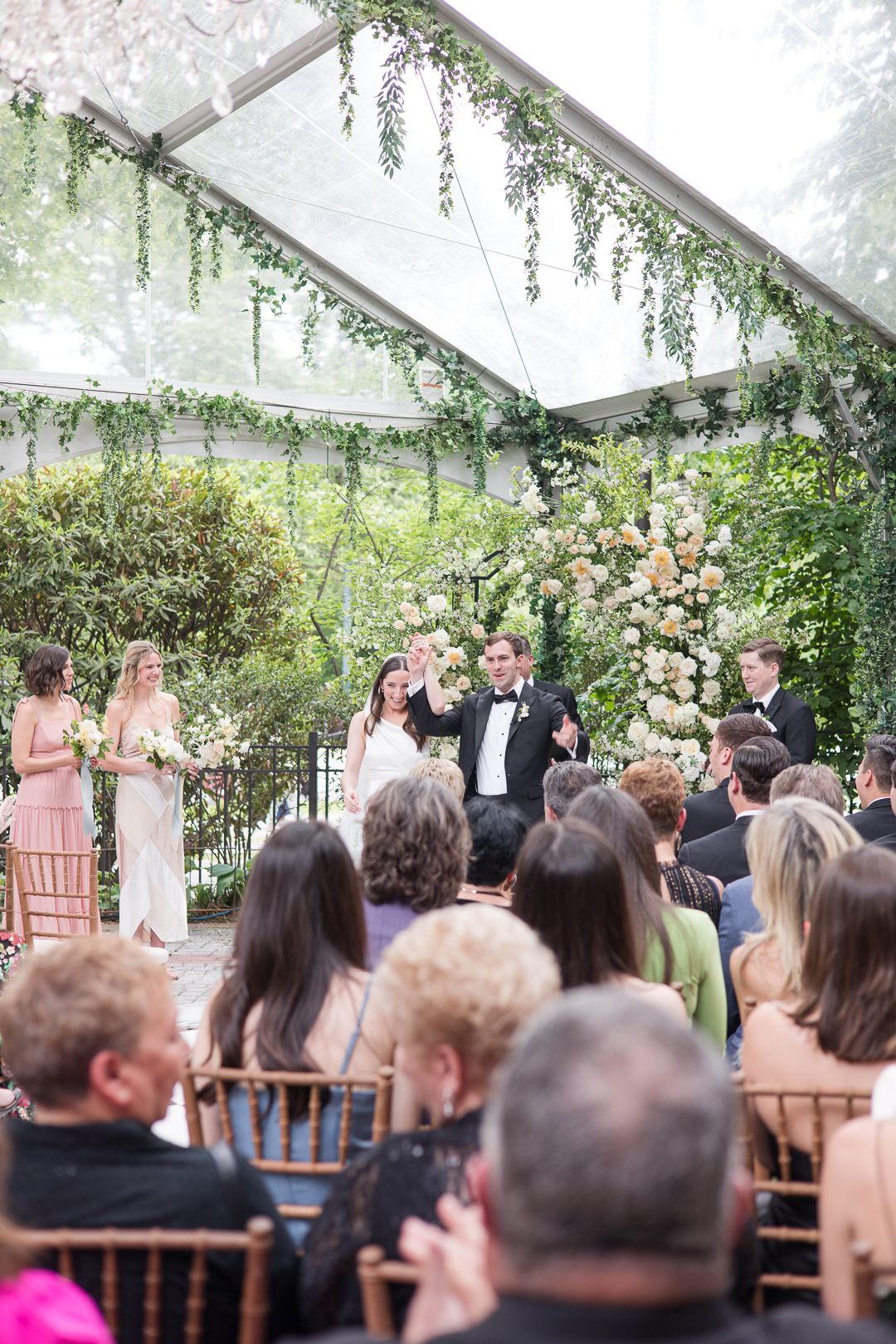 Wedding ceremony in progress under clear tent decorated with hanging greenery and floral arrangements.