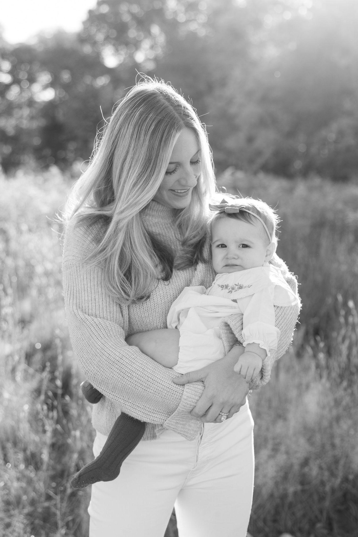 A tender outdoor portrait showing a parent and baby embracing in soft natural light against a blurred field background.