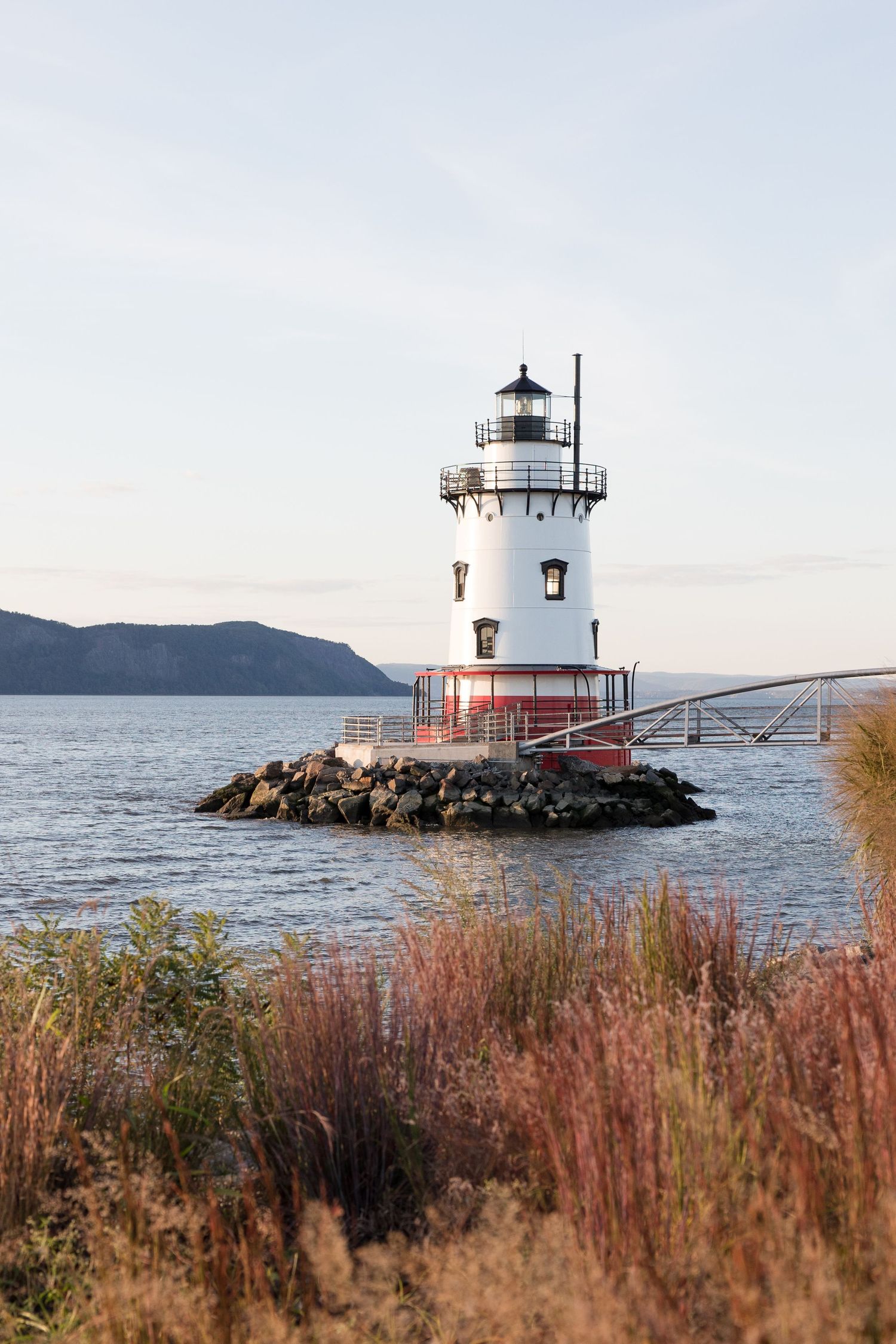 White and red lighthouse on rocky jetty with mountains and water in background.