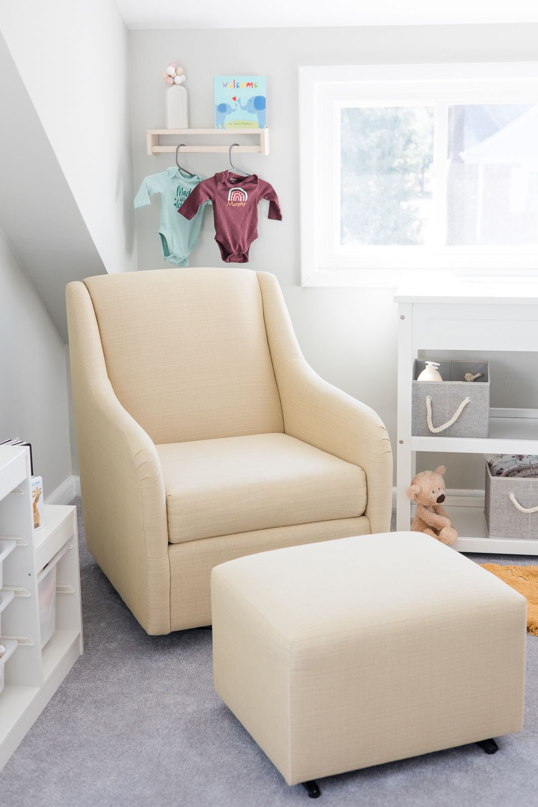 Cozy cream-colored glider chair and ottoman in a nursery with baby clothes and storage baskets.