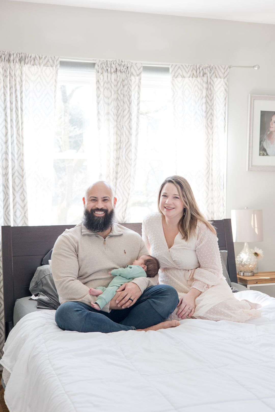 Family sits together on bed near windows with white patterned curtains.