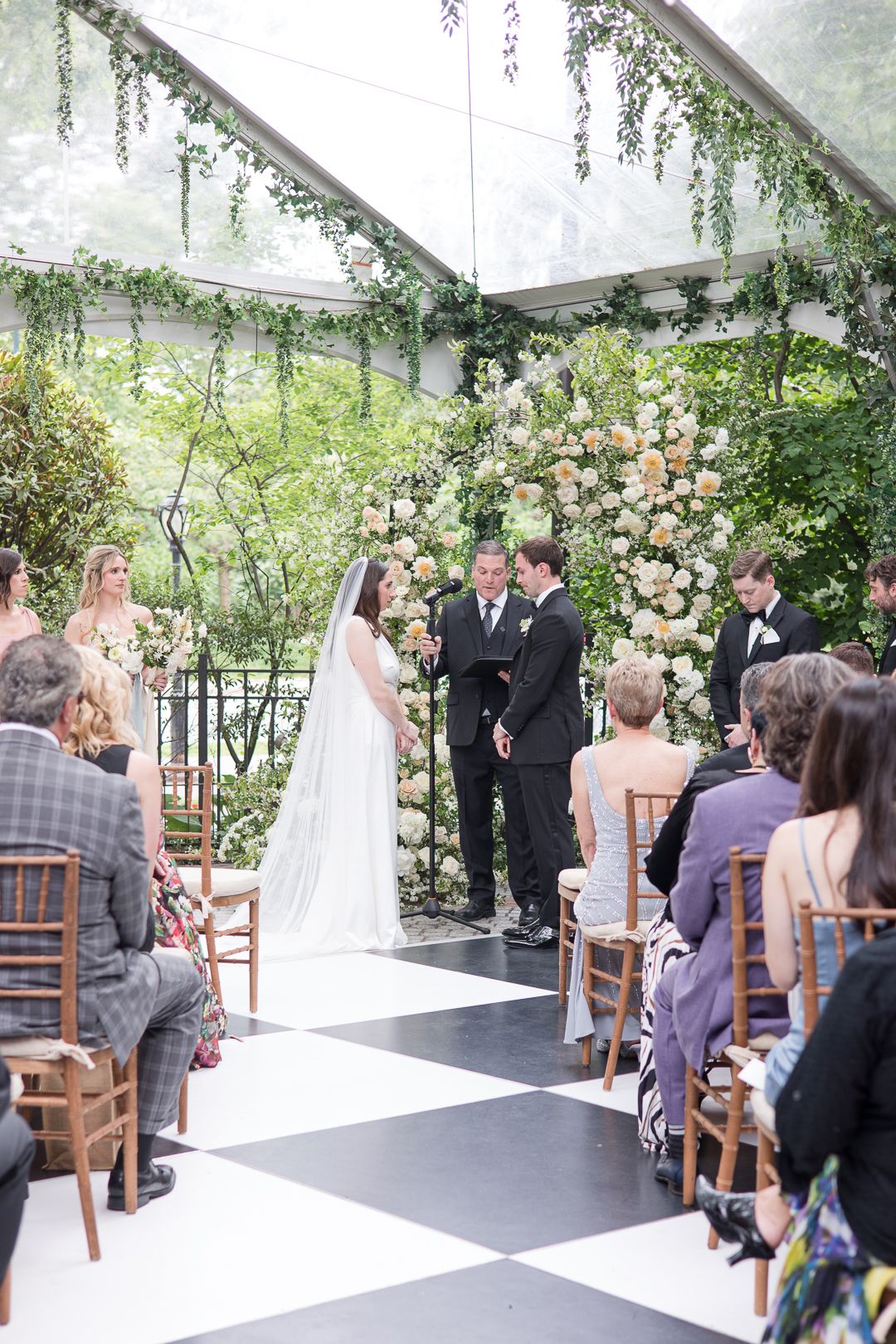 Indoor garden wedding ceremony under clear tent with floral arch and black and white checkered floor.
